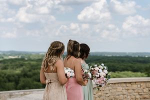 Three bridesmaids in pastel gowns stand together overlooking a scenic green landscape with dramatic clouds above.