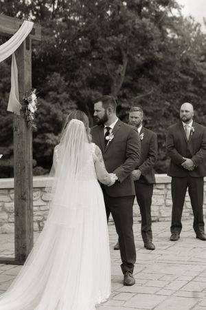 Black and white photo of bride and groom holding hands at outdoor wedding ceremony altar.