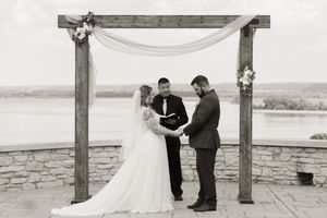 Black and white photo of bride and groom exchanging vows under wooden arch by waterfront.