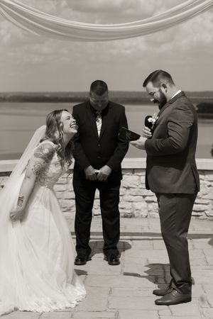 Black and white photo of laughing bride and groom exchanging vows with officiant at outdoor waterfront ceremony.