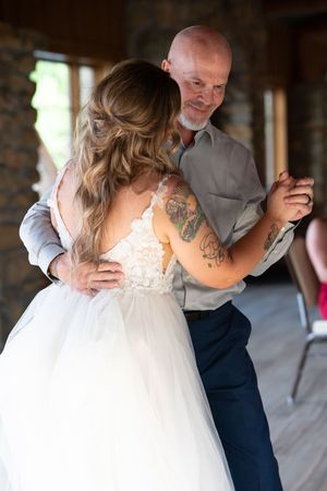 Emotional father-daughter dance at wedding reception with tattooed bride in white gown.