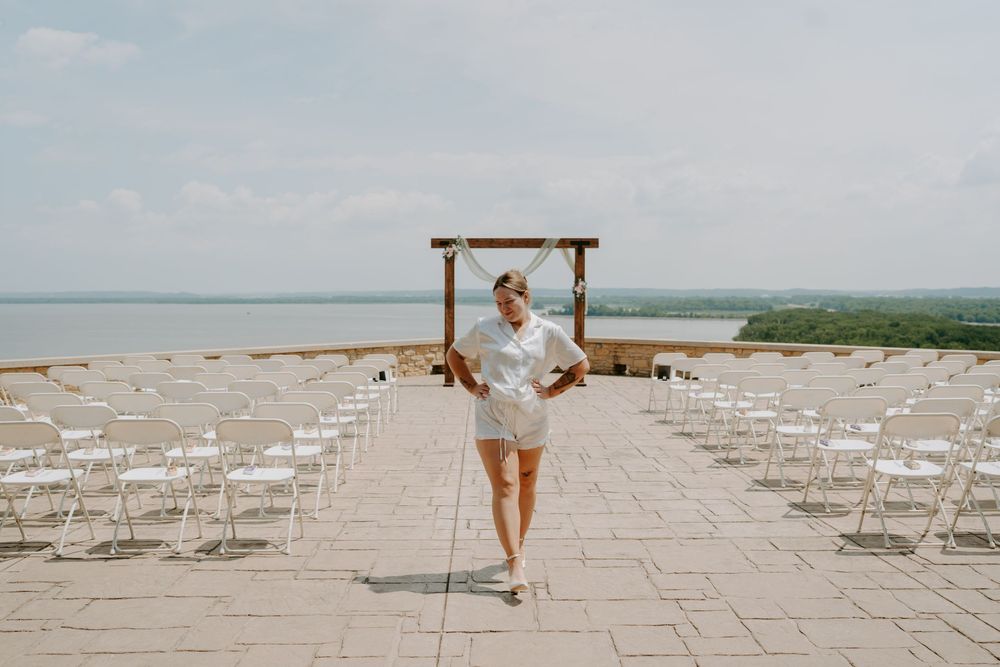 Woman in white outfit walks between rows of white chairs on stone terrace with wooden arch and lake view.