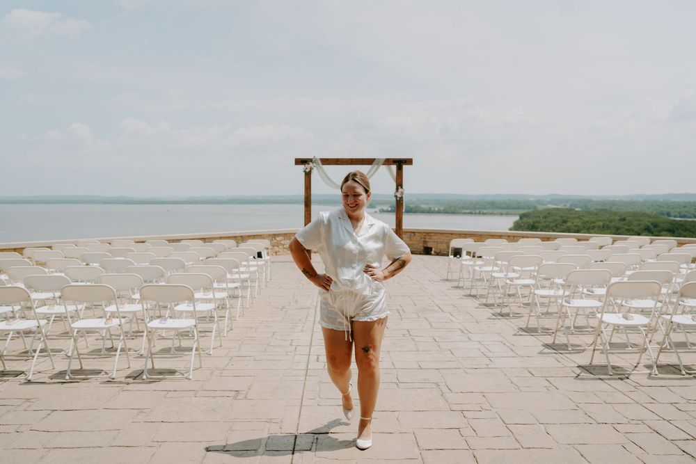 Wedding coordinator in white outfit standing confidently at outdoor ceremony venue overlooking scenic lake.