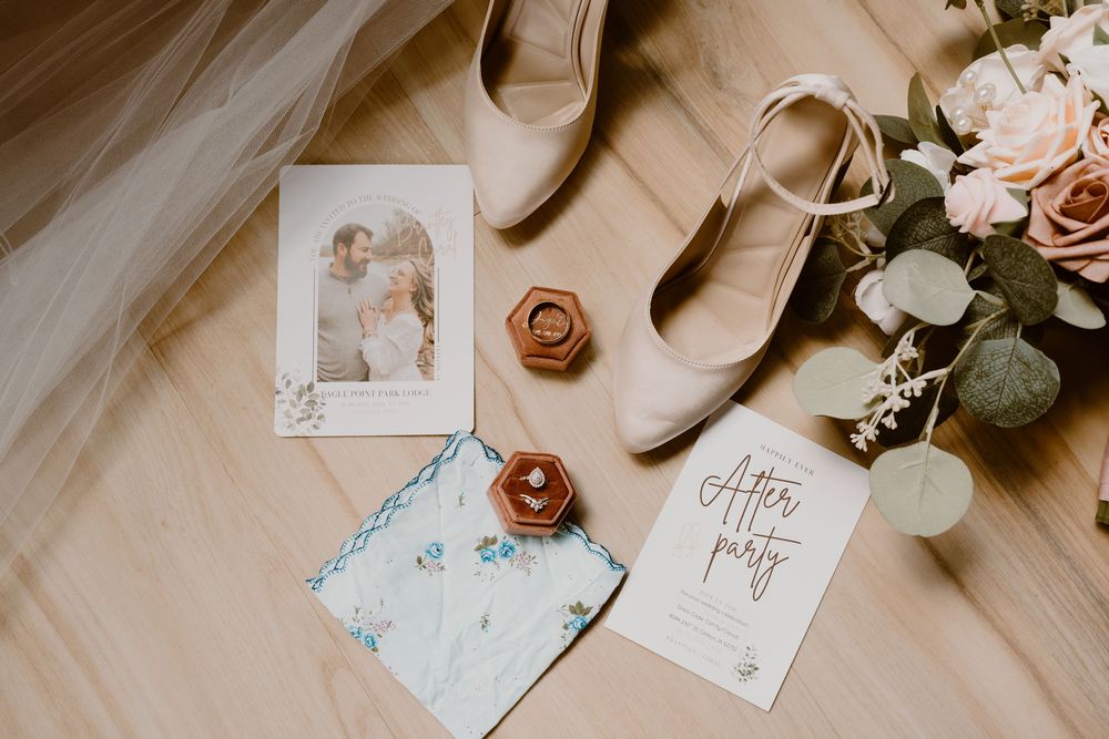 Wedding flat lay featuring invitation, rings, nude heels, floral bouquet, and after party card on wood floor.