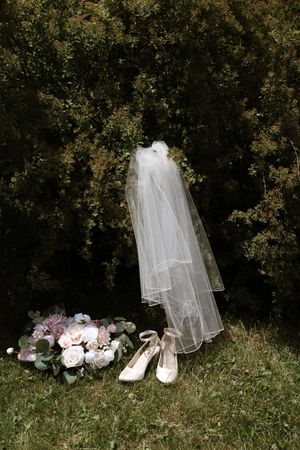 Bridal accessories displayed on grass including white veil, heels, and pink rose bouquet against dark green bushes.