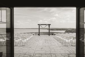 Black and white photo of an outdoor wedding ceremony setup with white chairs facing a wooden arch by the water.