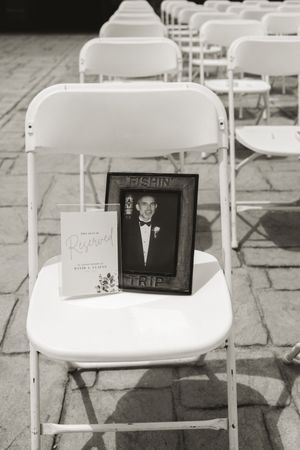 Black and white memorial photo of loved one displayed on white chair at outdoor wedding ceremony.