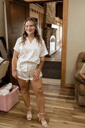 Woman in white satin shorts set and heels stands smiling in rustic wood-interior room with stone fireplace.