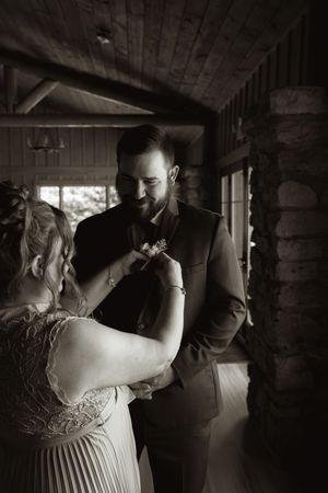 Black and white photo of bride pinning boutonniere on smiling groom inside rustic log cabin venue.