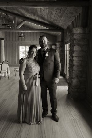 Black and white portrait of groom in suit with boutonniere posing with woman in formal gown inside rustic venue.