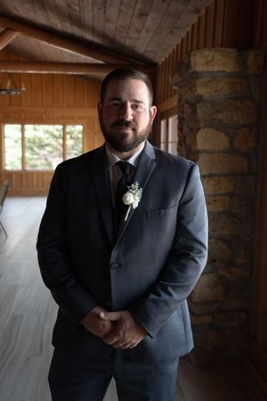 Handsome groom in a gray suit with boutonniere poses inside a rustic wooden wedding venue.