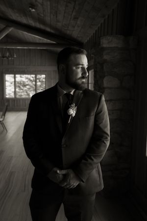 Black and white portrait of bearded groom in suit with boutonniere by rustic window.