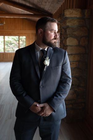 Bearded groom in gray suit with boutonniere gazing out window in rustic wood venue.