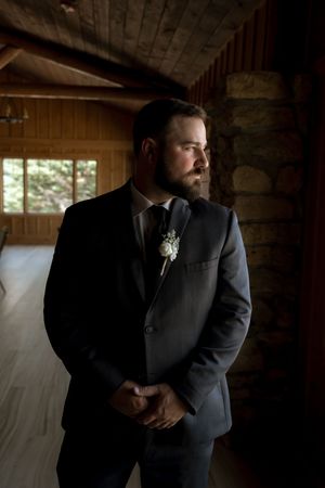 Groom in dark suit with white boutonniere standing thoughtfully in rustic wooden venue with stone wall.