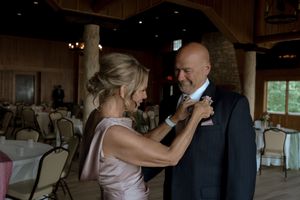 Woman pins boutonniere on mans lapel inside rustic wooden wedding venue reception hall.