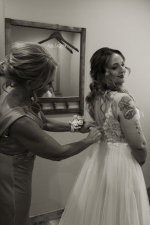 Black and white photo of woman helping bride fasten lace wedding gown, mirror reflection visible in background.