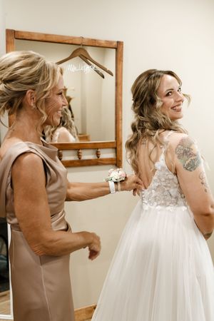 Mother helping bride button her lace wedding gown while smiling in a bridal suite mirror.