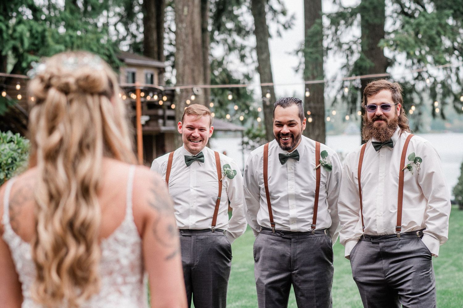 Three groomsmen in suspenders and bow ties smile at a bride during an outdoor wedding ceremony among tall trees.