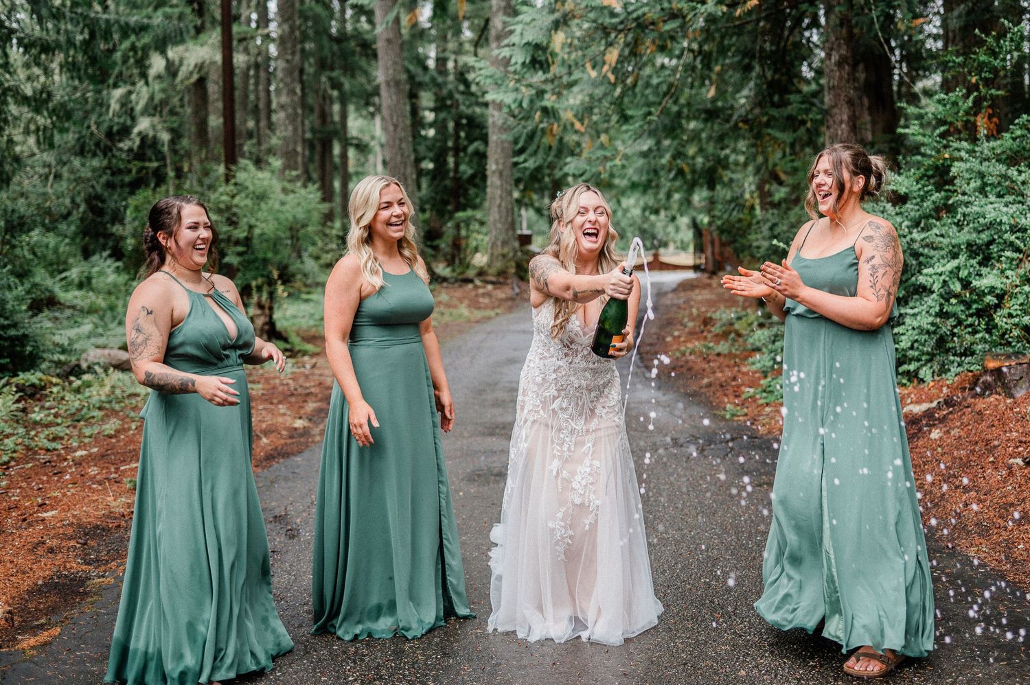 Bride pops champagne with three bridesmaids in sage green gowns, laughing outdoors on a forest path.