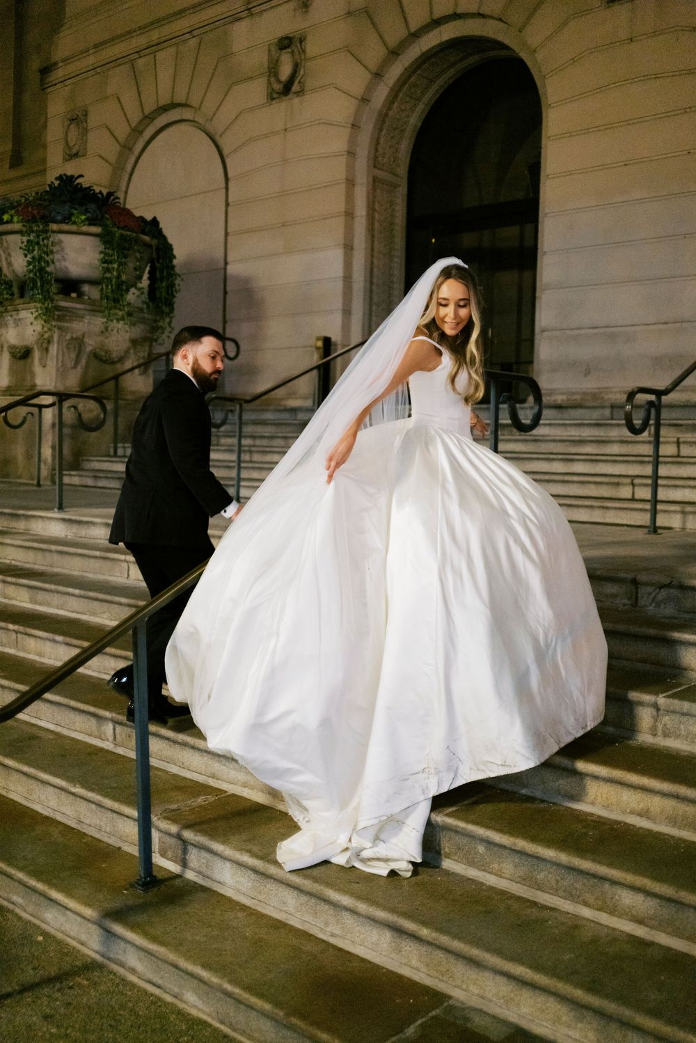 Bride in flowing white gown and veil descends stone steps with groom in black tuxedo at Chicago Art Institute.