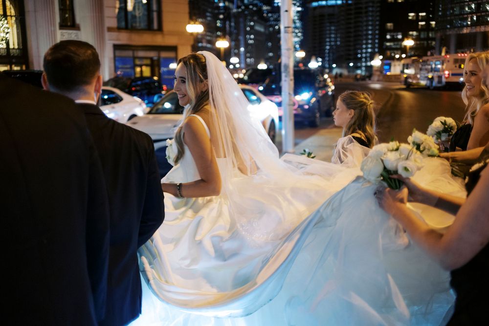 Bride in elegant white ball gown with veil being assisted by wedding party on cinematic Chicago street at night.