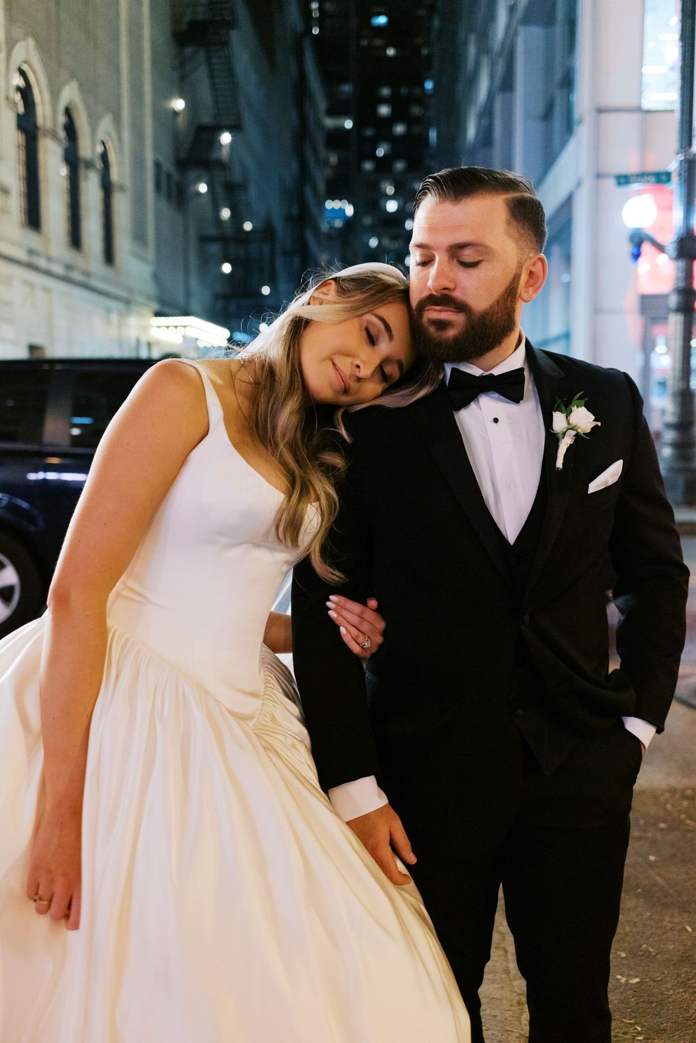 Elegant bride and groom in black tuxedo sharing romantic moment on Chicago city street at night.