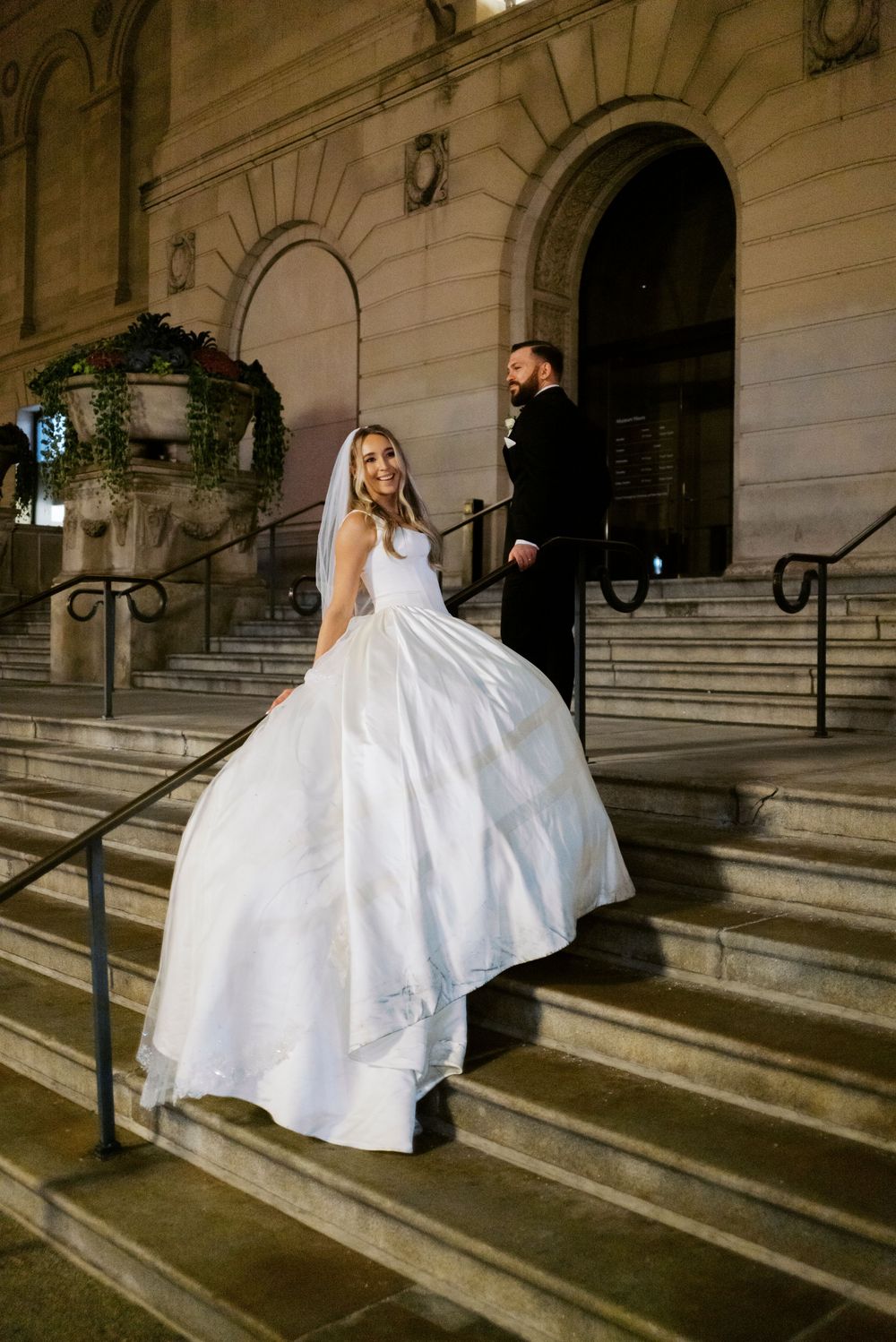 Bride in white ball gown and veil poses on Art Institute of Chicago's stone steps with groom in black tuxedo at night.