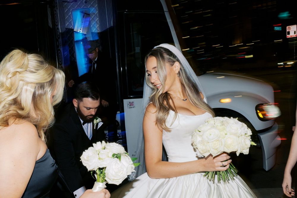 Bride in strapless wedding gown holding white bouquet arriving at wedding venue beside party bus at night.