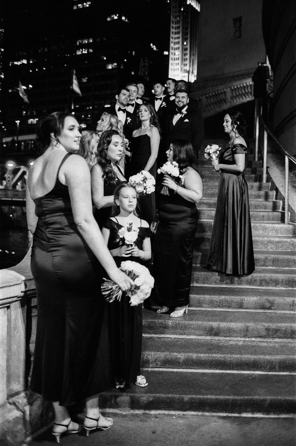 Wedding party in black formal attire poses on Chicago Riverwalk stone steps at night holding white bouquets.