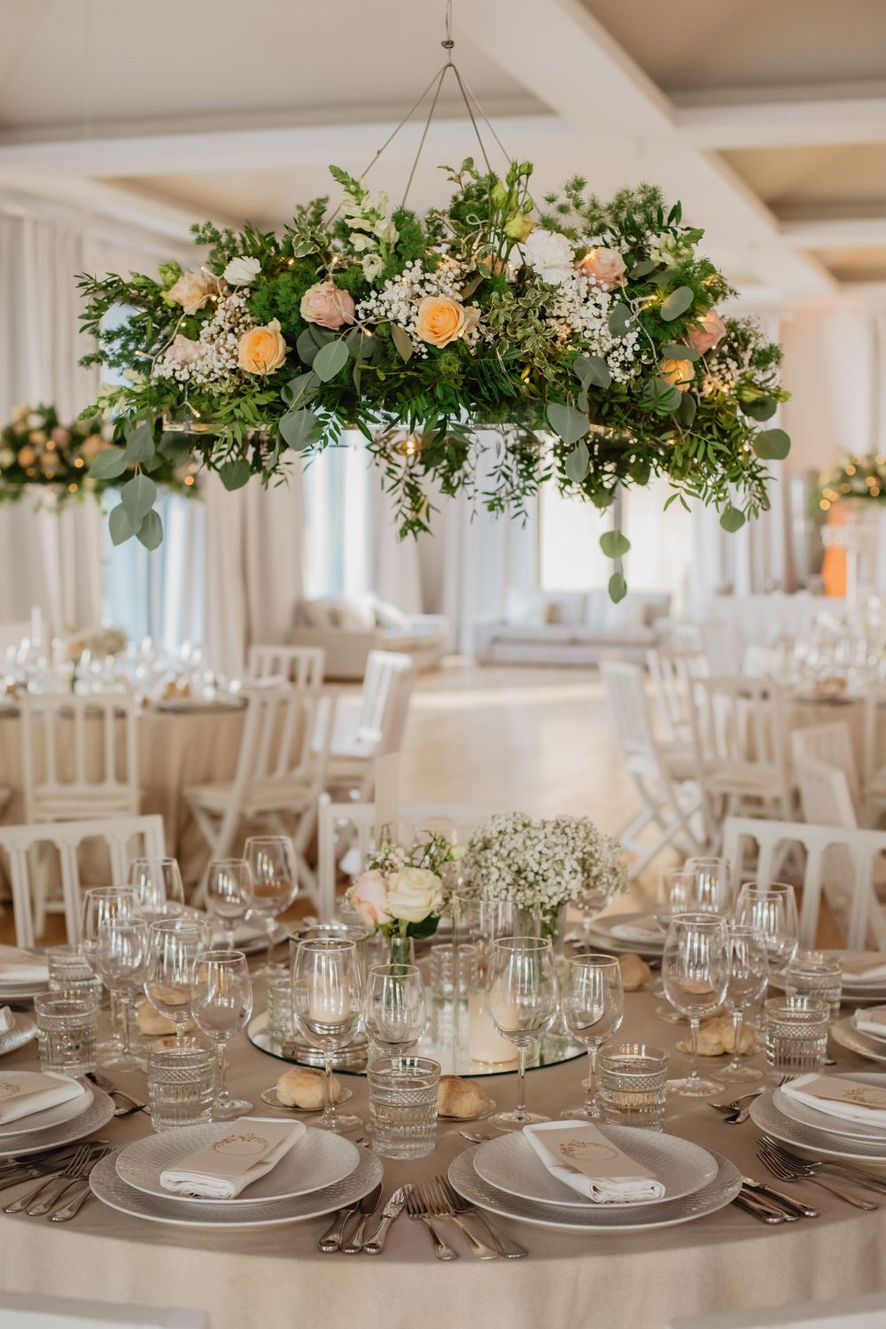 Elegant wedding reception table with hanging floral centerpiece featuring roses, eucalyptus, and greenery in a bright venue.