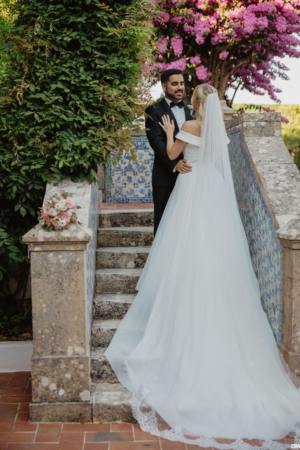 Bride in white gown with veil and groom in tuxedo embrace on stone steps surrounded by lush greenery and pink flowers.