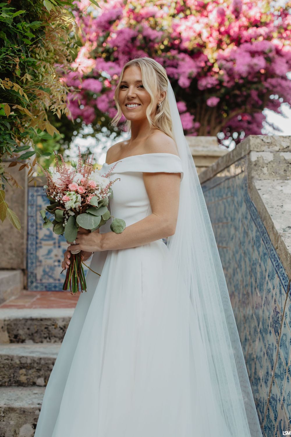 Smiling bride in off-shoulder white gown holding bouquet with pink flowers and veil outdoors.