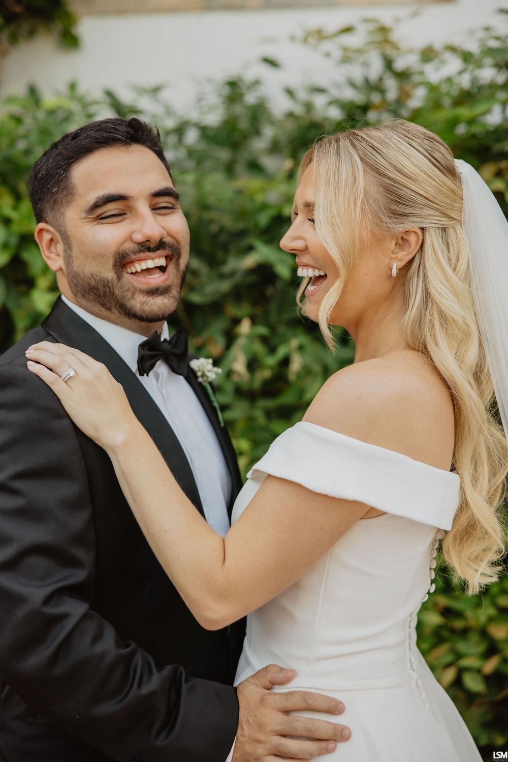 Laughing bride and groom in black tuxedo embracing outdoors on wedding day.