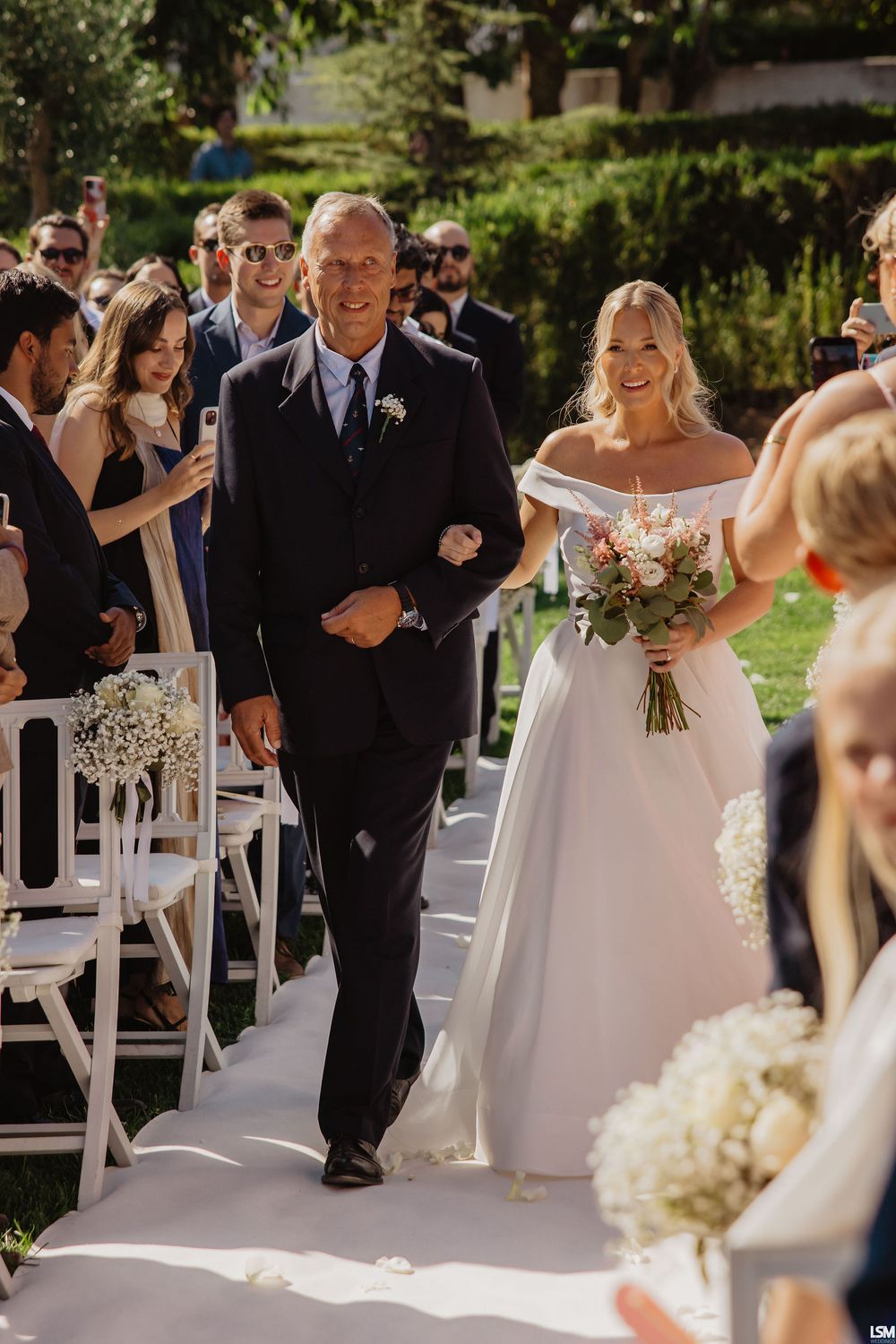Bride in off-shoulder gown walks down outdoor aisle with escort, holding floral bouquet, guests seated on white chairs.