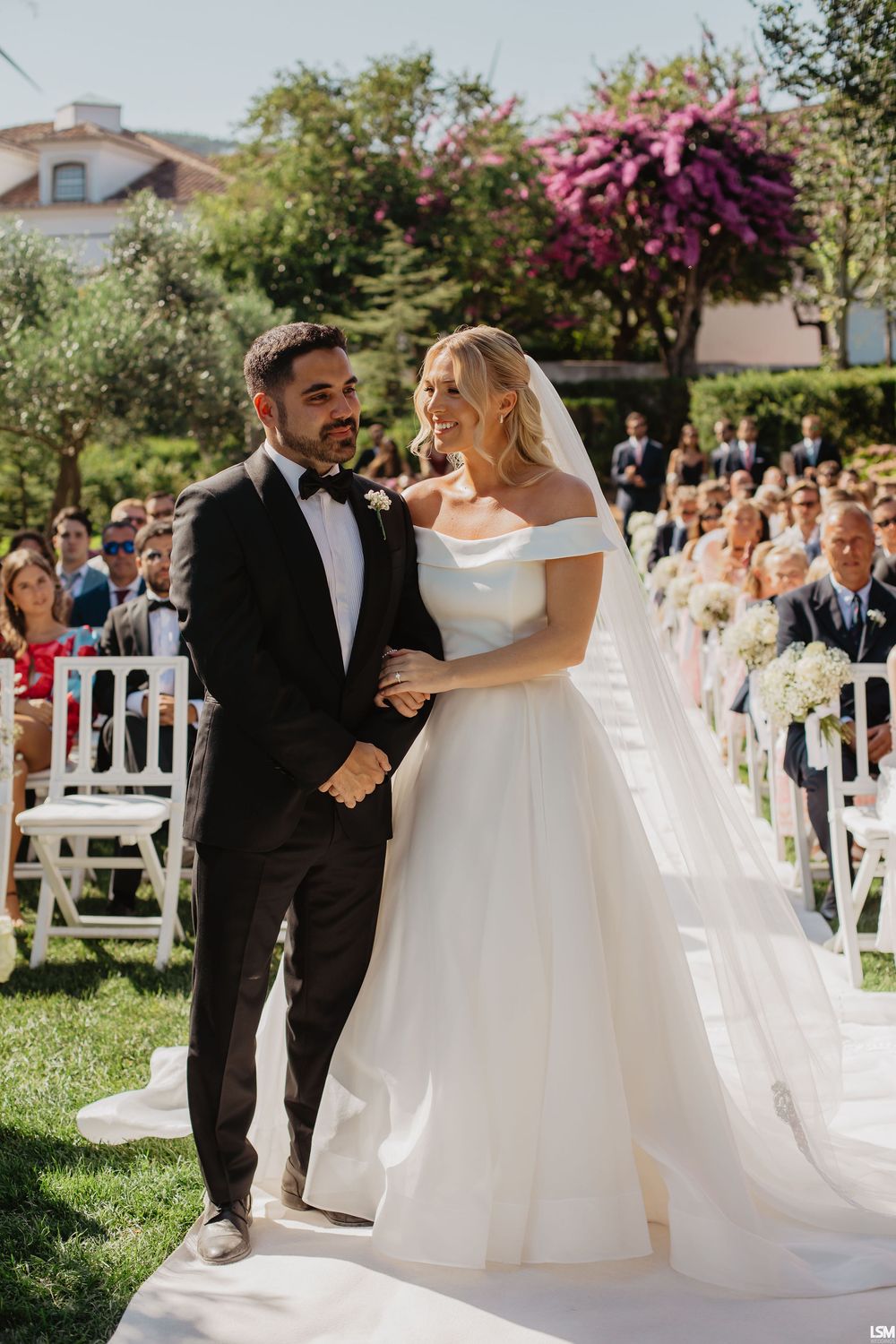 Bride in off-shoulder ball gown and groom in black tuxedo walking down outdoor aisle at garden wedding ceremony.