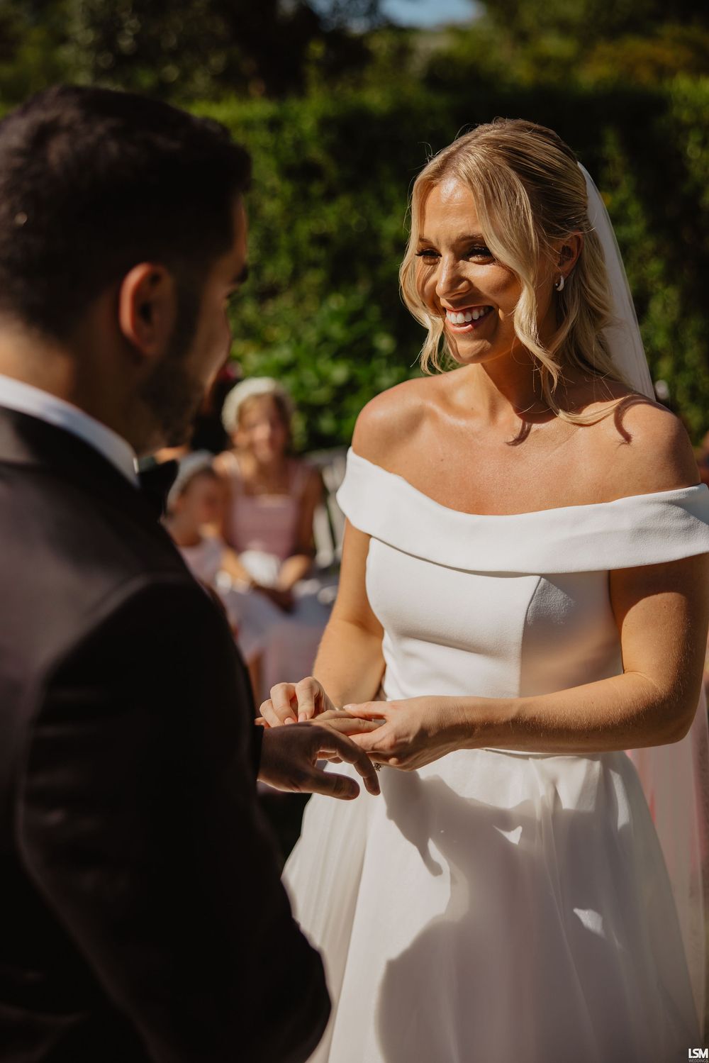 Smiling bride in off-shoulder white gown exchanges rings with groom during outdoor wedding ceremony.