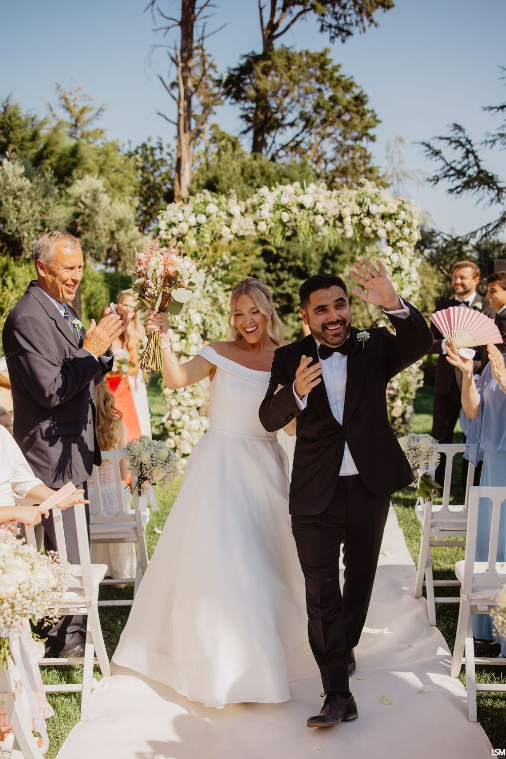 Joyful newlyweds celebrate recessional at outdoor garden wedding ceremony with floral arch and cheering guests.