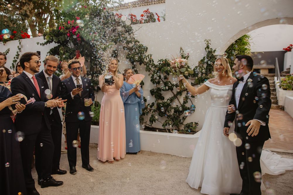 Bride and groom celebrate with bubbles surrounded by smiling wedding guests in elegant attire at an outdoor venue.