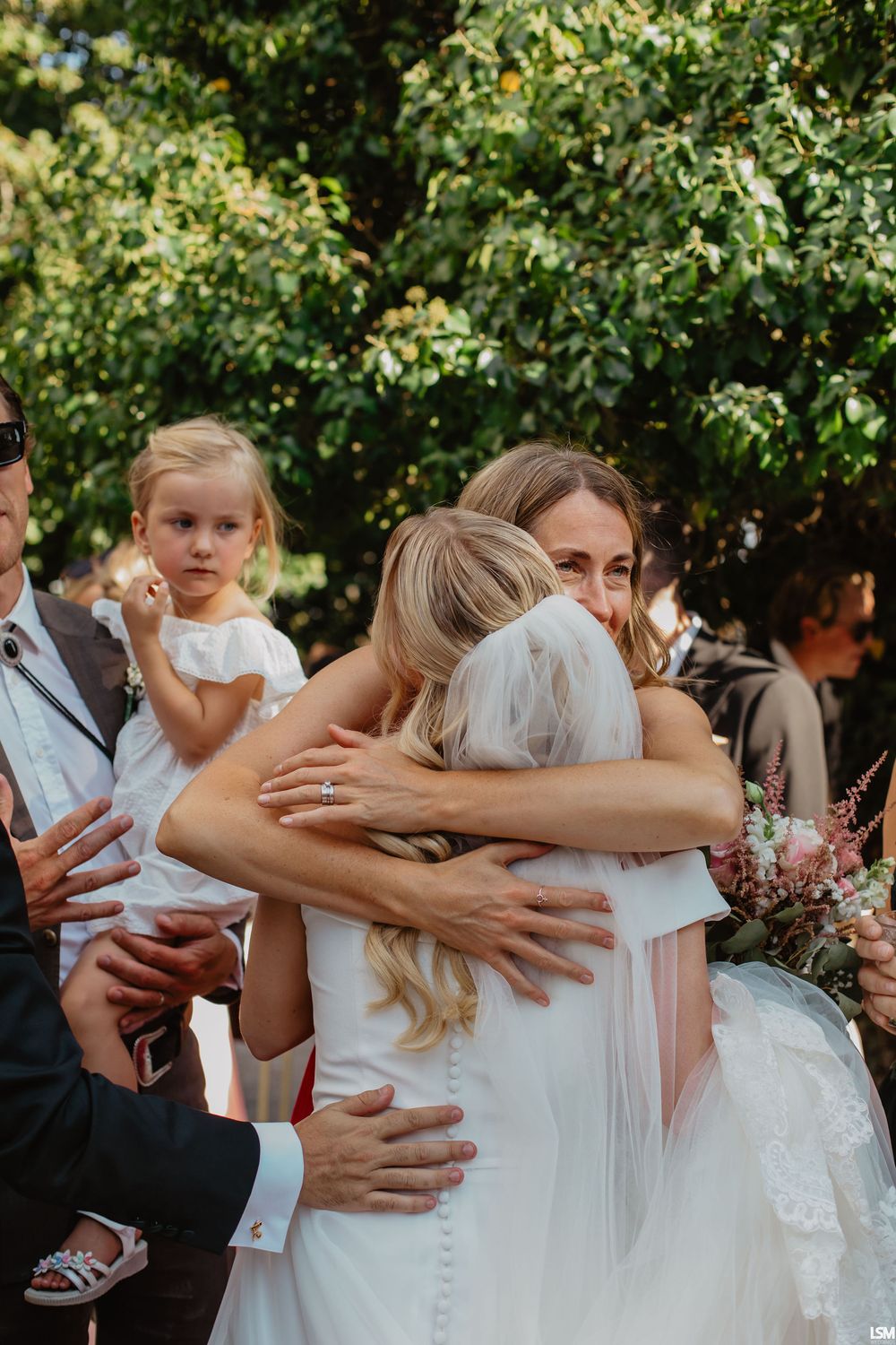 Bride in white gown and veil shares emotional hug with guest at outdoor wedding ceremony surrounded by greenery.