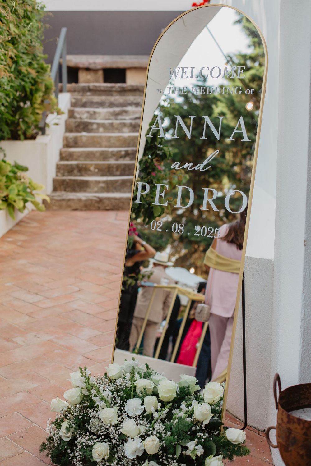 Gold mirror wedding welcome sign for Anna and Pedro dated 02.08.2025, adorned with white roses and greenery.
