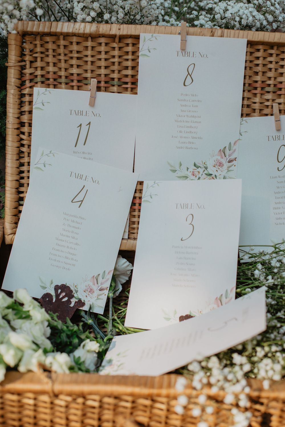 Elegant wedding seating chart cards with floral designs displayed on a wicker backdrop at an outdoor reception.