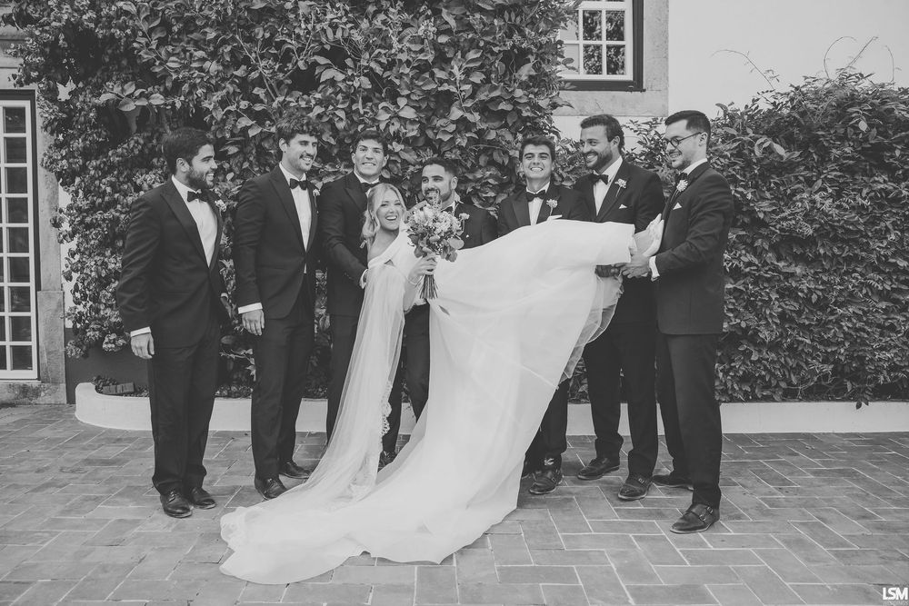 Black and white wedding photo of groom carrying bride surrounded by smiling groomsmen in tuxedos outdoors.