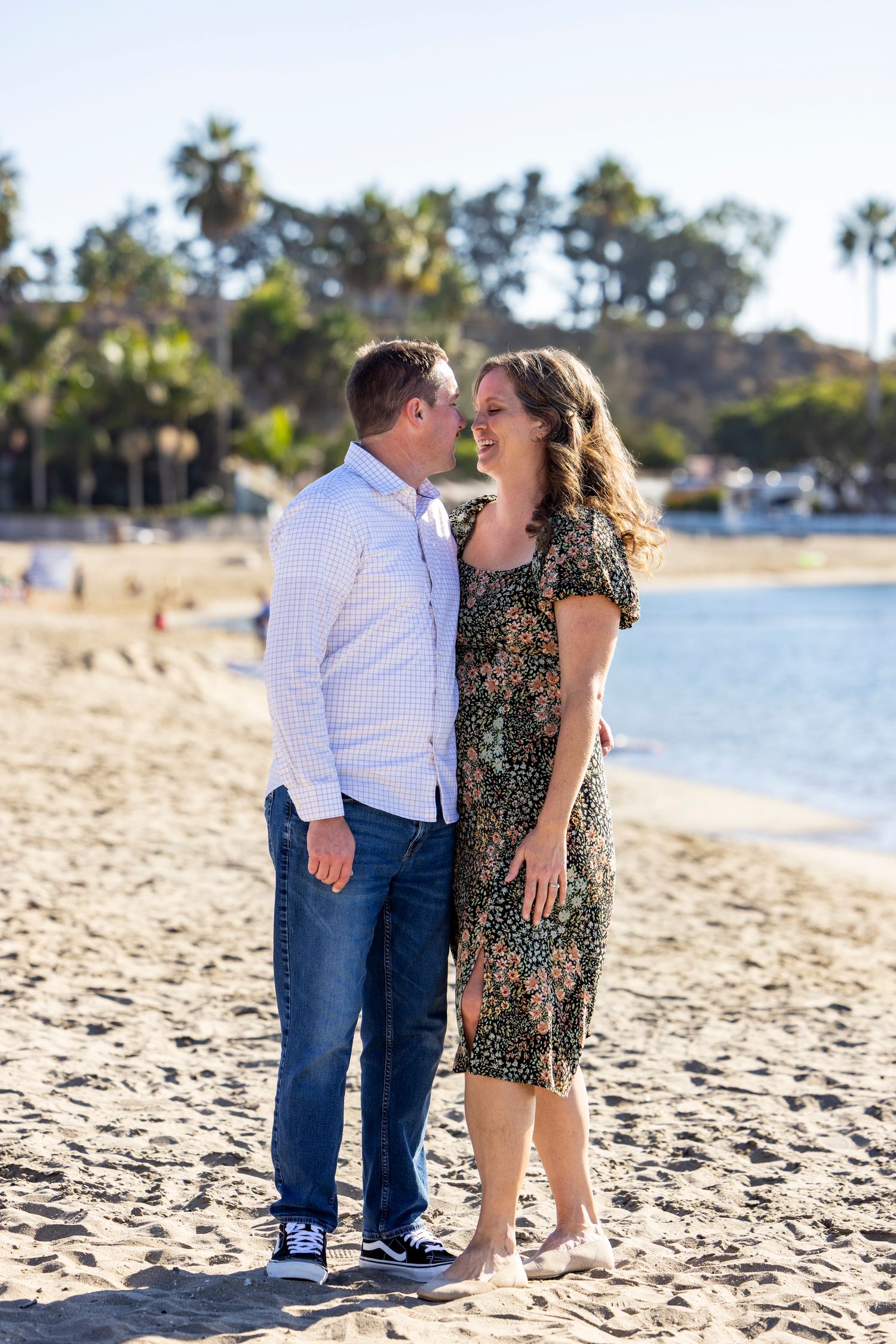 Couple sharing a romantic moment on a sunny beach, man in white shirt and jeans, woman in floral dress, palm trees behind.