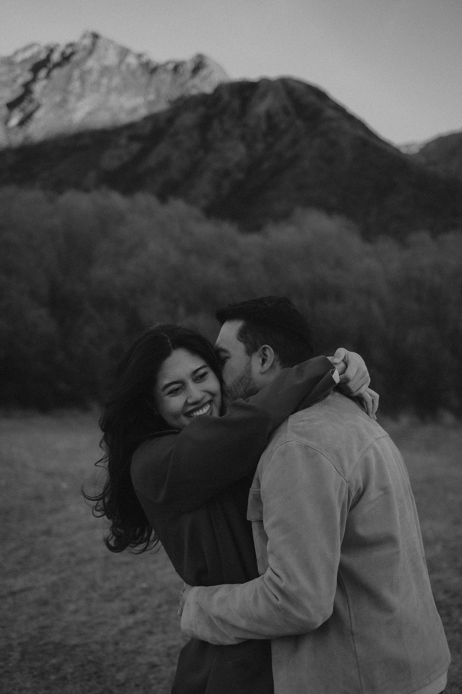 Black and white photo of a laughing couple embracing with a snow-capped mountain and trees in the background.