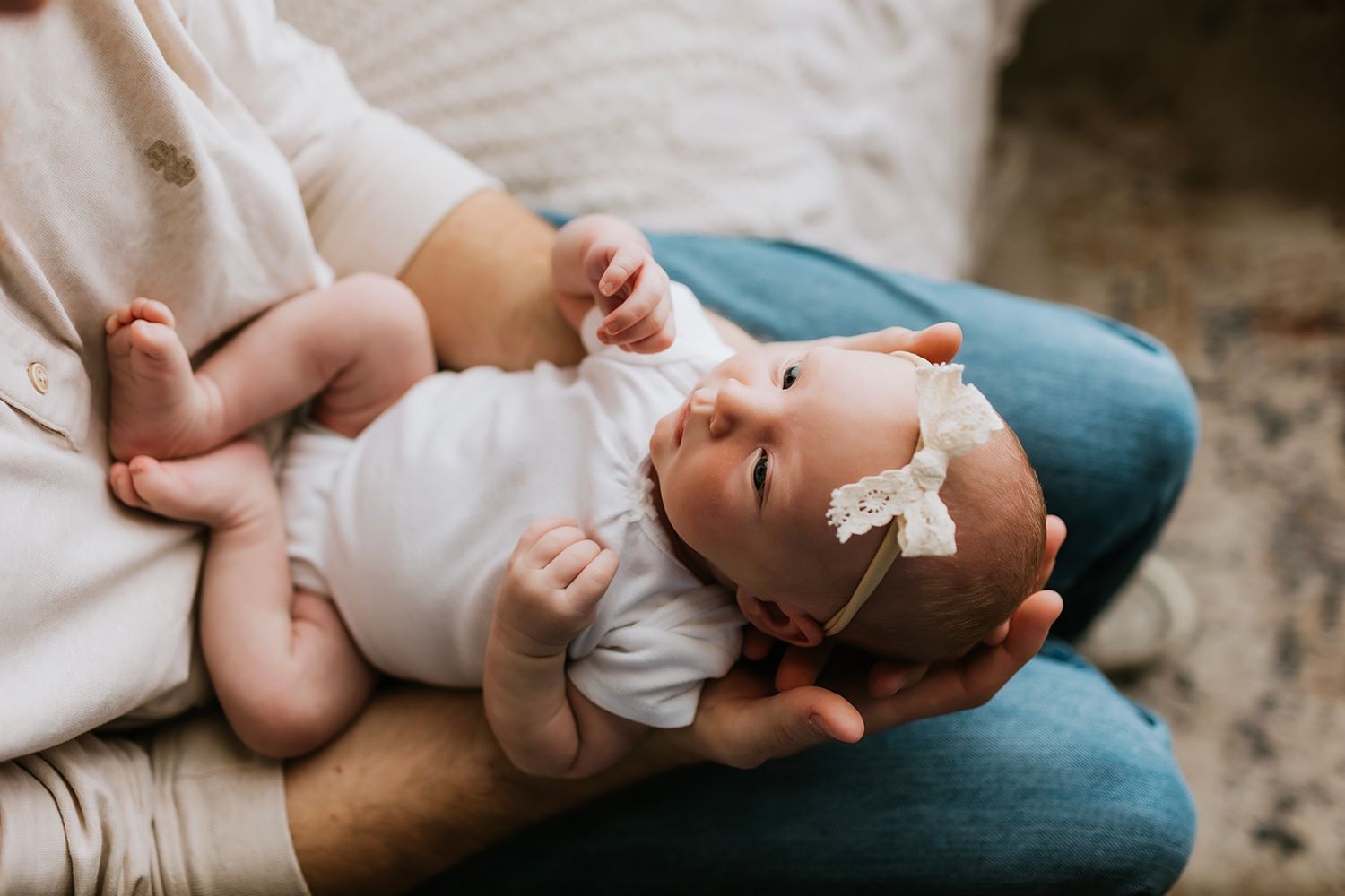 A newborn baby girl wearing a white onesie and floral headband rests peacefully in a parent's arms.