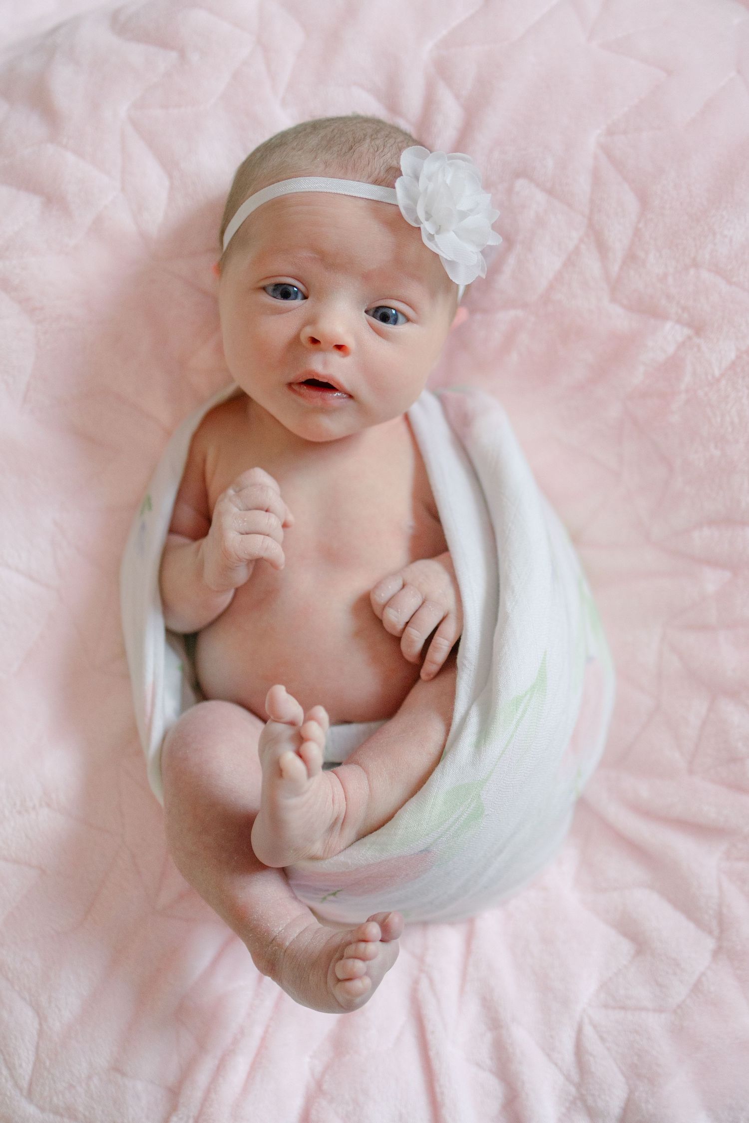 Newborn baby girl with blue eyes and white flower headband wrapped in swaddle blanket on pink quilt.