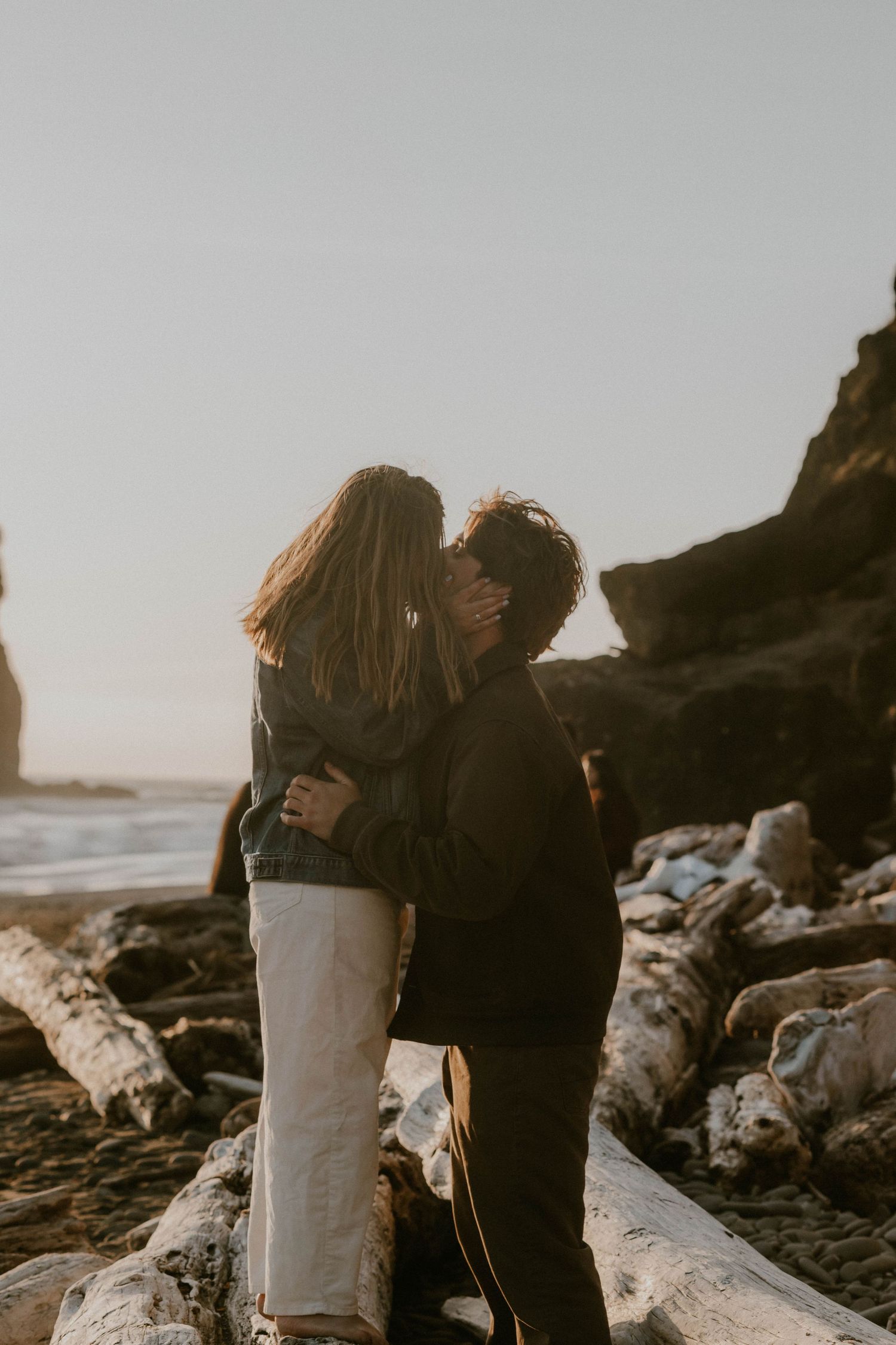 A couple shares a tender kiss outdoors at golden hour, with trees and blue sky in the background.