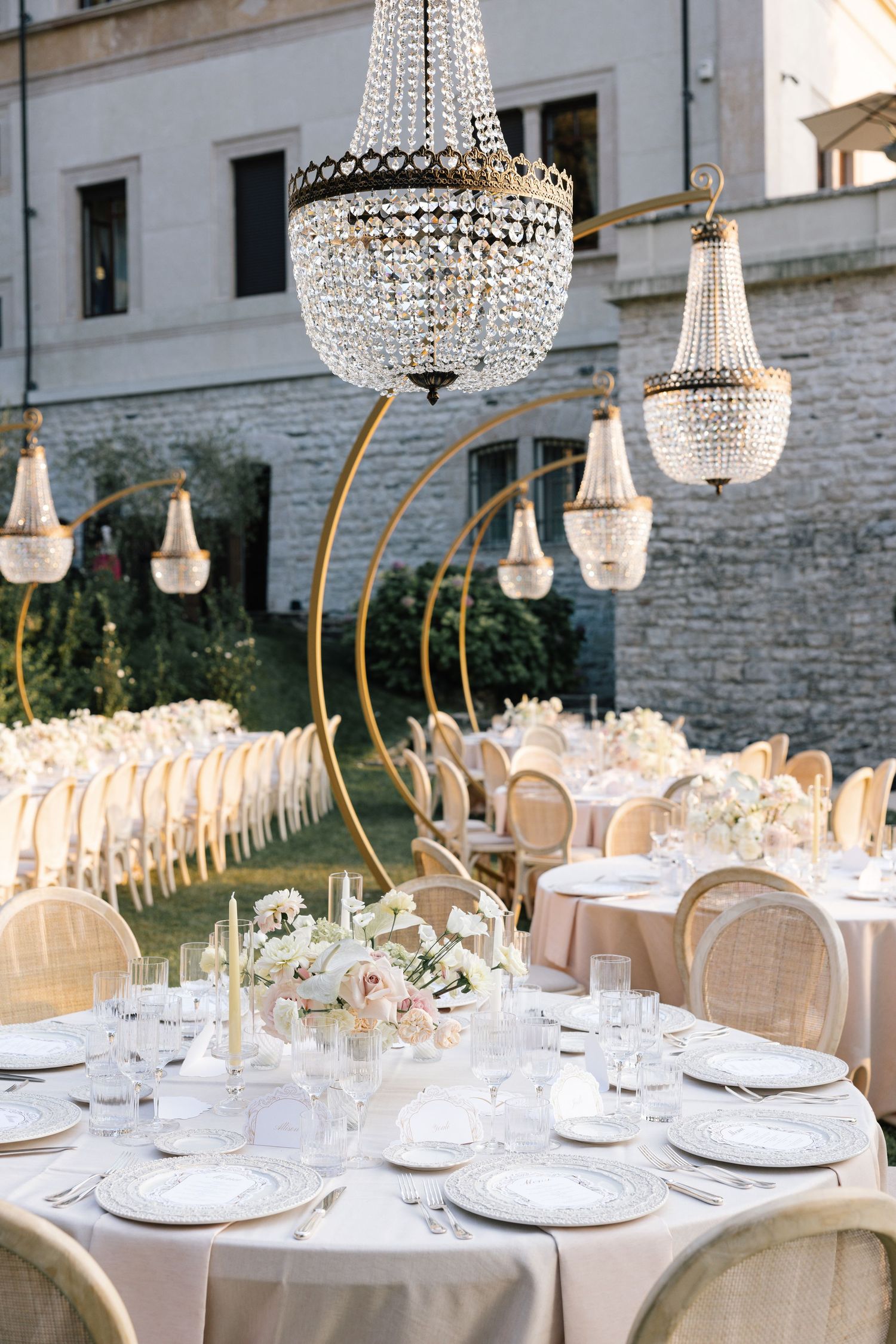 A couple walks down an outdoor aisle lined with flowers and cypress trees leading to an elegant villa entrance.