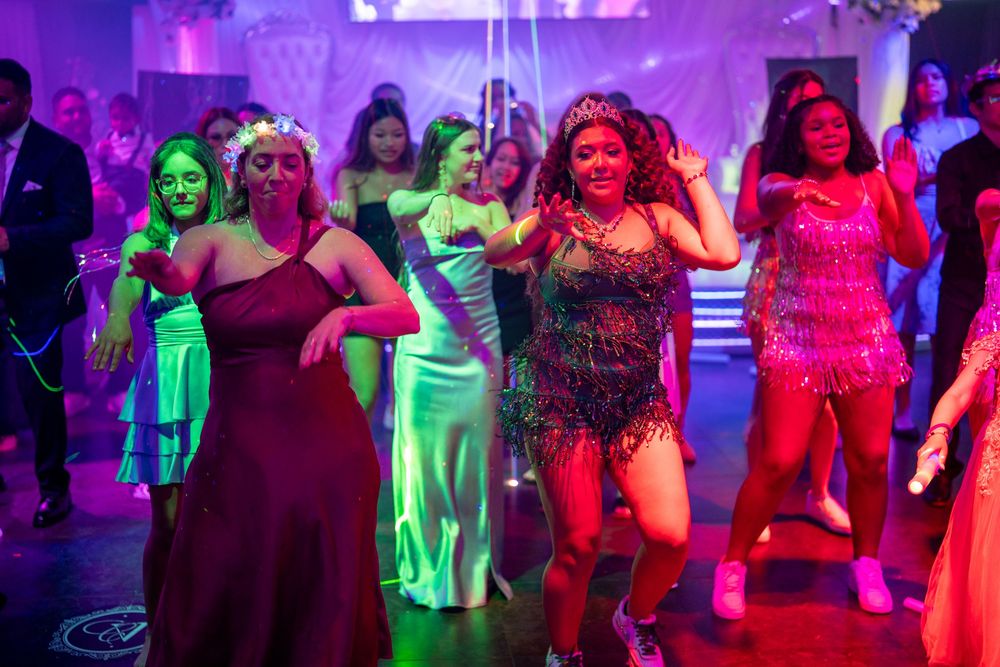 Women dancing energetically at a glamorous prom or formal event under colorful purple and green lights.