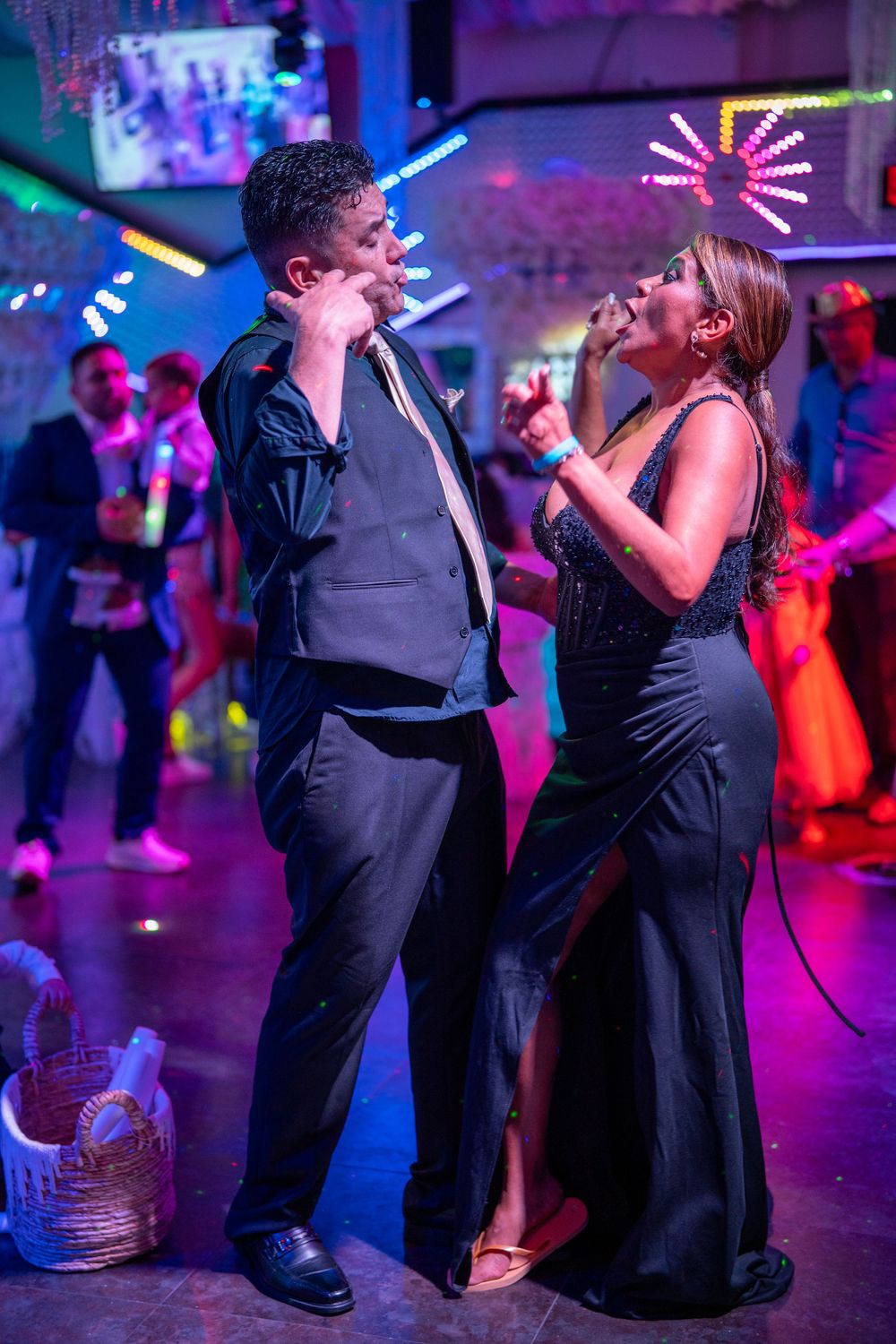 Couple dancing elegantly at a vibrant nightclub, man in black suit and woman in black gown under colorful neon lights.